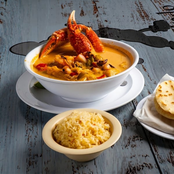 A white bowl of seafood soup with crab claws, rice, and flatbread on a rustic blue table.