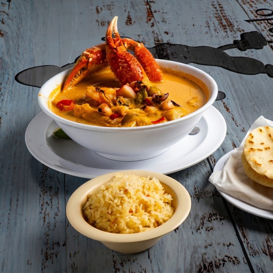 A white bowl of seafood soup with crab claws, rice, and flatbread on a rustic blue table.