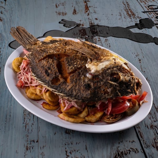Whole fried fish with plantains, pickled onions, and tomatoes on a white oval plate.