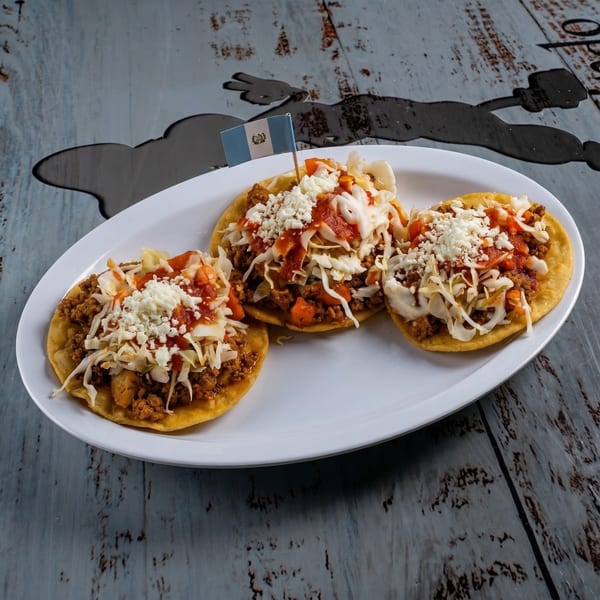 Three Guatemalan tostadas with meat, cabbage, sauce, and cheese on a white plate with a flag.